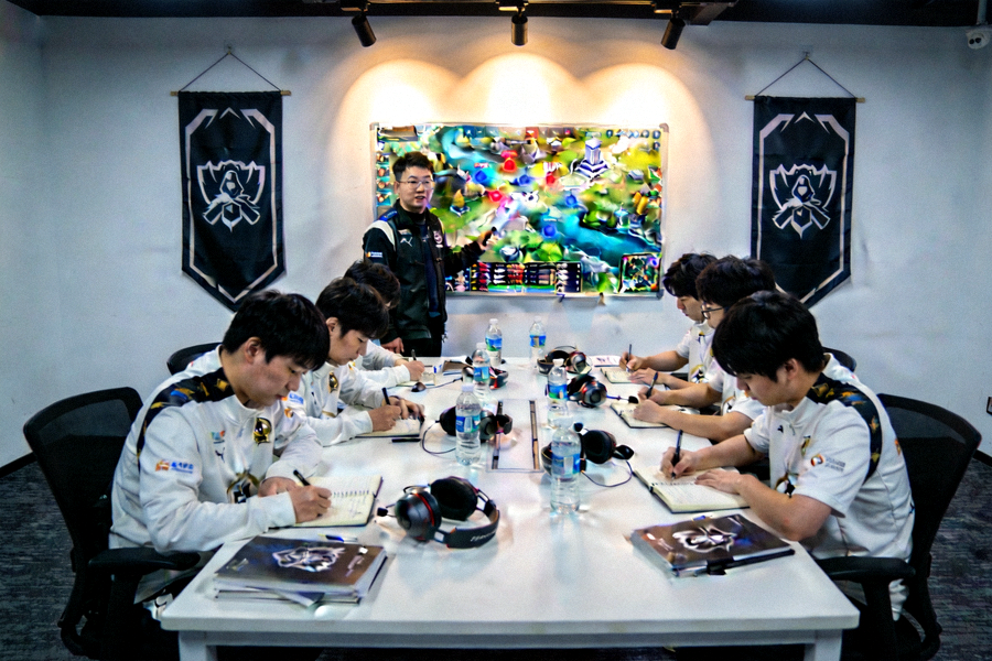 职业战队复盘会议场景，教练在战术板前讲解地图资源分配与视野布控策略，队员专注记录关键节点